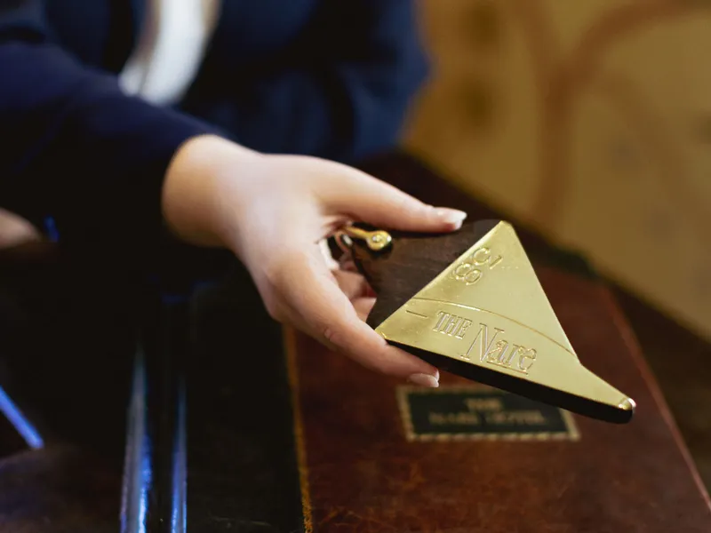 A hotel staff member holding a brass room key tag for The Nare above a leather-bound guest book.