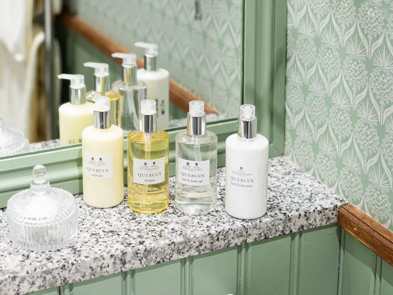 Bathroom sink with Quercus toiletries on a granite countertop beneath a mirror and extendable shaving mirror, set against pale green patterned wallpaper.