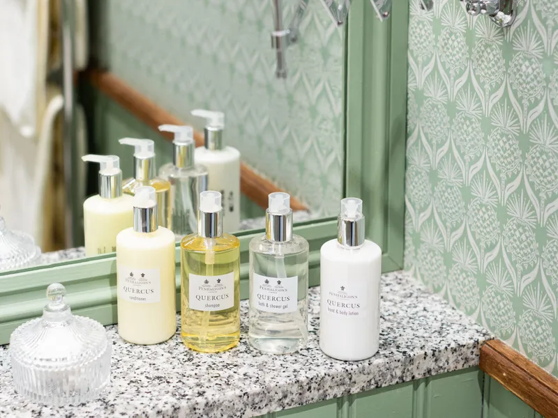 Bathroom sink with Quercus toiletries on a granite countertop beneath a mirror and extendable shaving mirror, set against pale green patterned wallpaper.