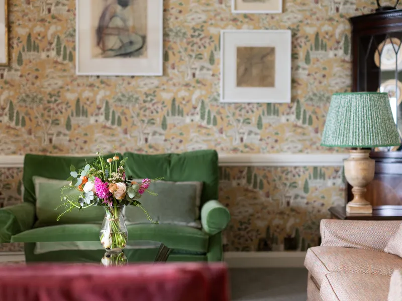 Elegant sitting room with patterned wallpaper, a green sofa, framed artwork, a floral arrangement on a glass table, and a lamp beside a dark wooden cabinet