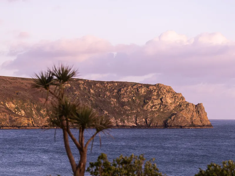 Swimming pool overlooking a calm bay and rocky headland at sunset, with soft pink clouds overhead