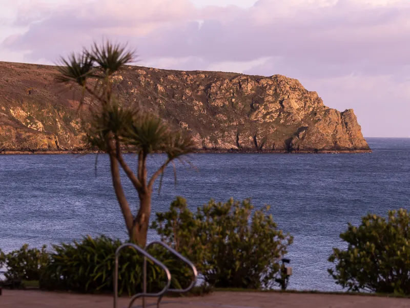 Swimming pool overlooking a calm bay and rocky headland at sunset, with soft pink clouds overhead