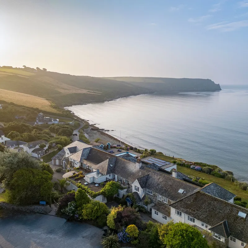 Aerial view of a coastal hotel and cottages beside a calm bay at sunrise, with rolling countryside and cliffs in the distance.