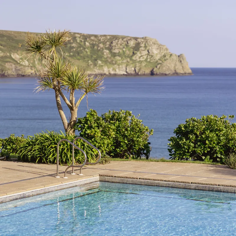 Outdoor swimming pool overlooking a calm blue sea with a rocky coastal headland in the distance.
