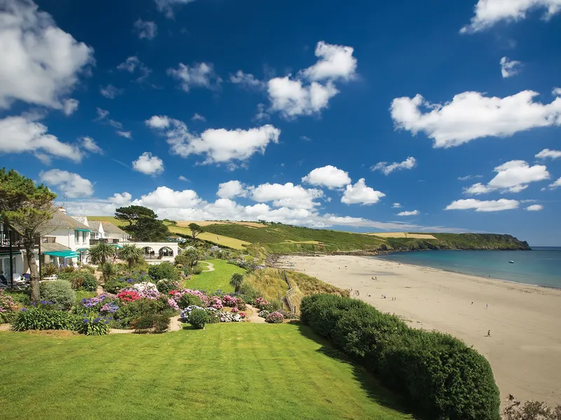 Coastal hotel and landscaped gardens overlooking a sandy beach and calm blue sea under a bright sky with scattered clouds