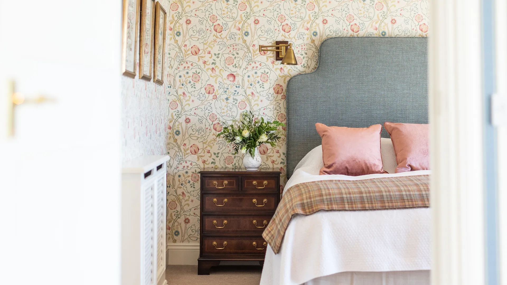 Elegant bedroom with floral wallpaper, grey upholstered headboard, pink cushions and a wooden bedside chest
