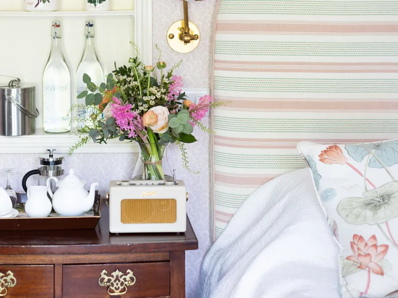 Cosy bedroom corner with a striped upholstered headboard, brass wall light, wooden chest of drawers and built-in shelves with books, jars and flowers.