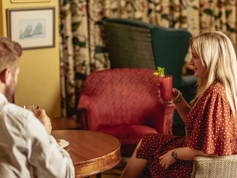 Woman holding a pink cocktail while seated across from a man in a cosy lounge