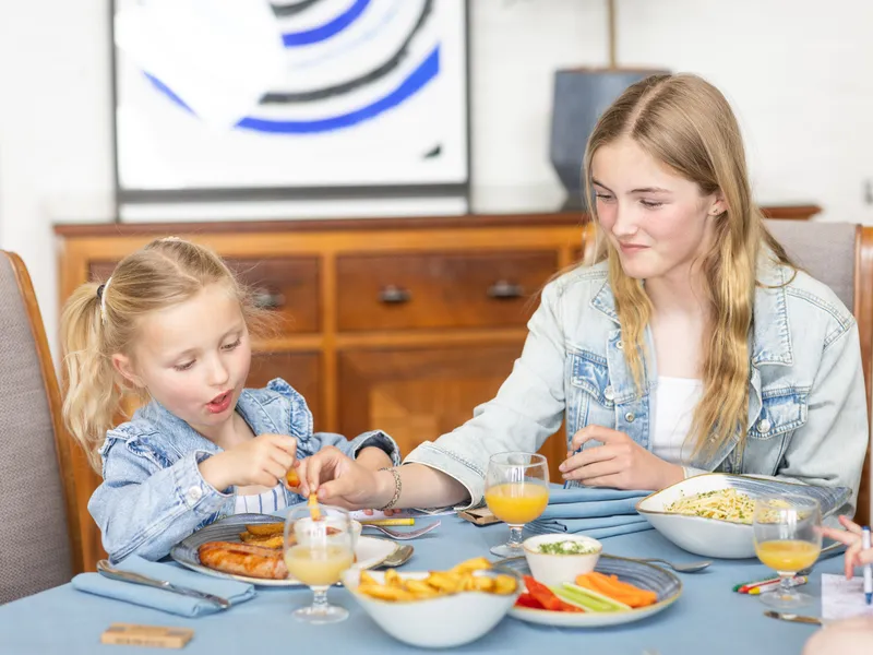 Two young girls in denim jackets sitting at a dining table, eating a meal with chips, vegetables and glasses of orange juice.