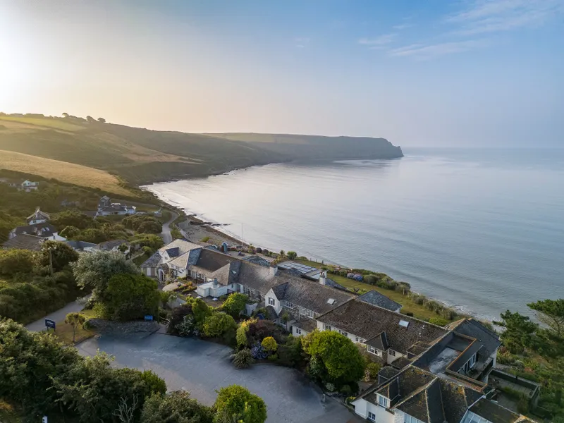 Aerial view of a coastal hotel and houses beside a calm bay with cliffs and countryside in soft evening light.