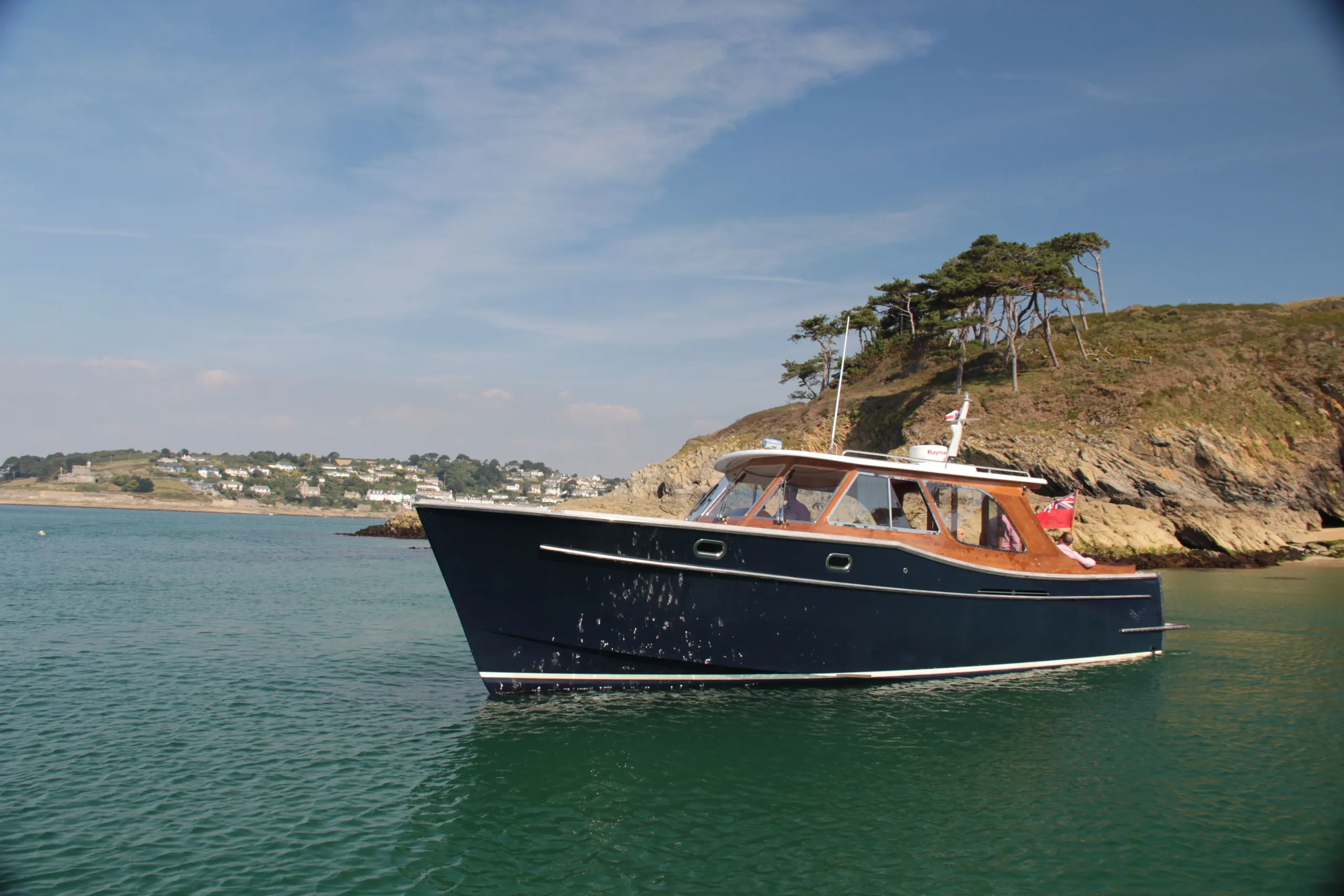 Dark blue motor boat anchored on calm coastal water near a rocky headland with pine trees
