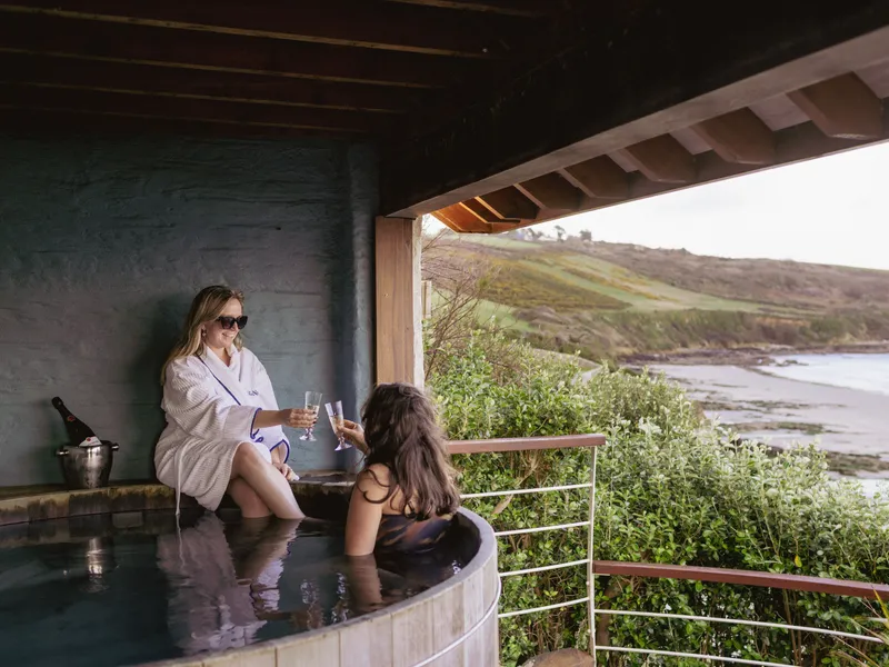 Two women relax in a covered outdoor hot tub overlooking a sandy bay and rolling coastal hills, raising glasses in a toast.