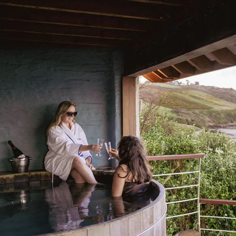 Two women relaxing in an outdoor hot tub with drinks, overlooking a quiet beach and rolling coastal hills.