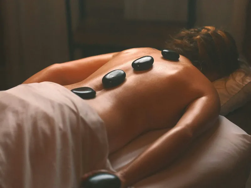 Person lying face down on a massage table with hot stones placed along their back in a dimly lit spa room.