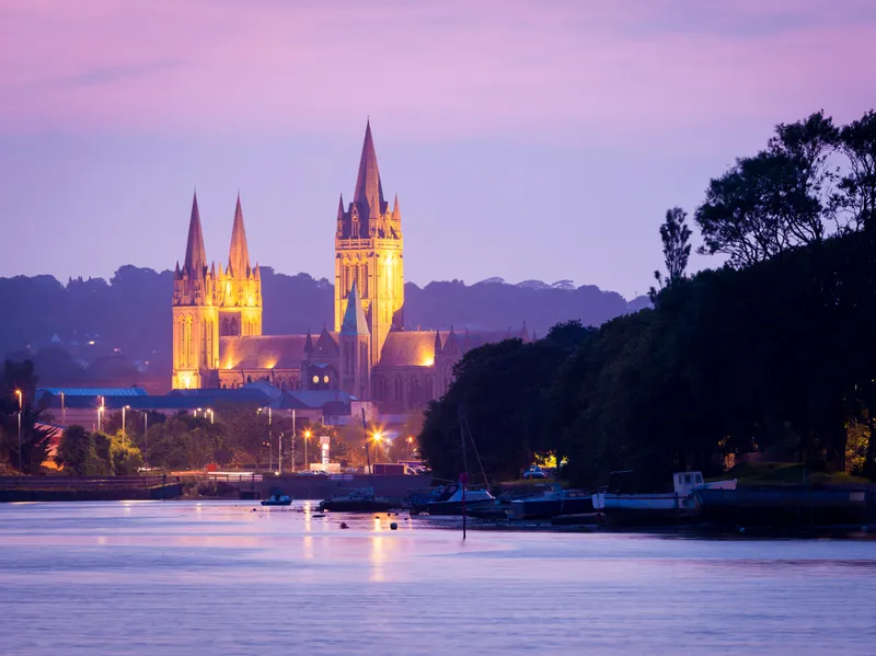 Floodlit Truro cathedral beside a river at dusk with boats in the foreground