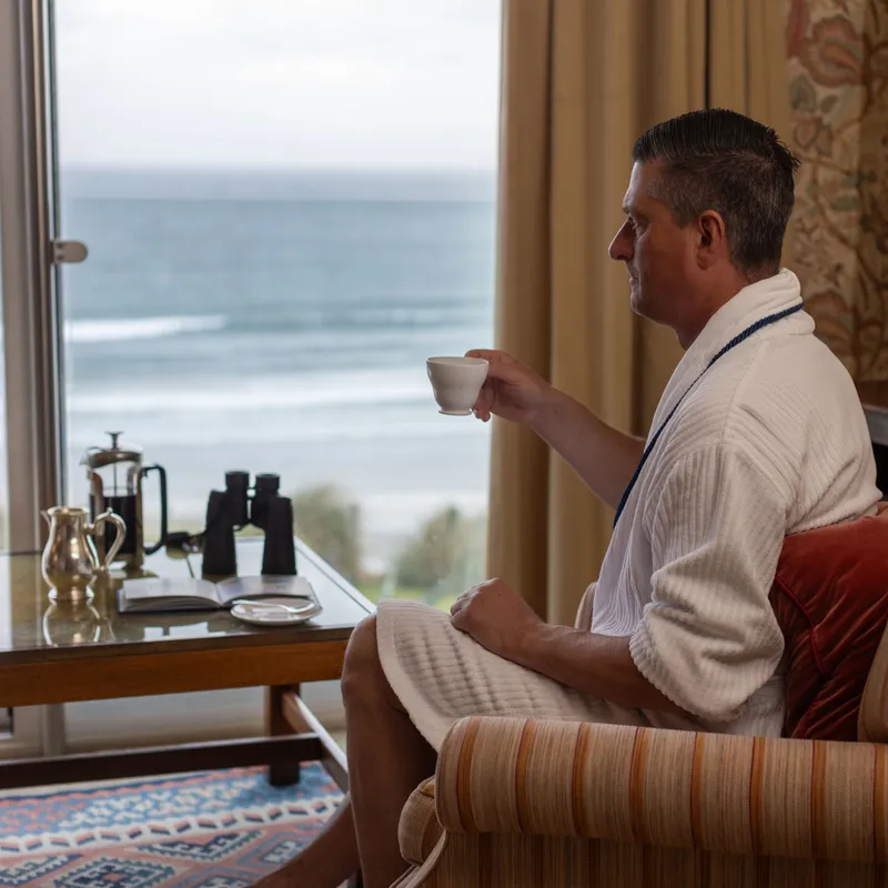 Man in a white dressing gown sitting by a seaside window, holding a cup of tea in a cosy hotel room