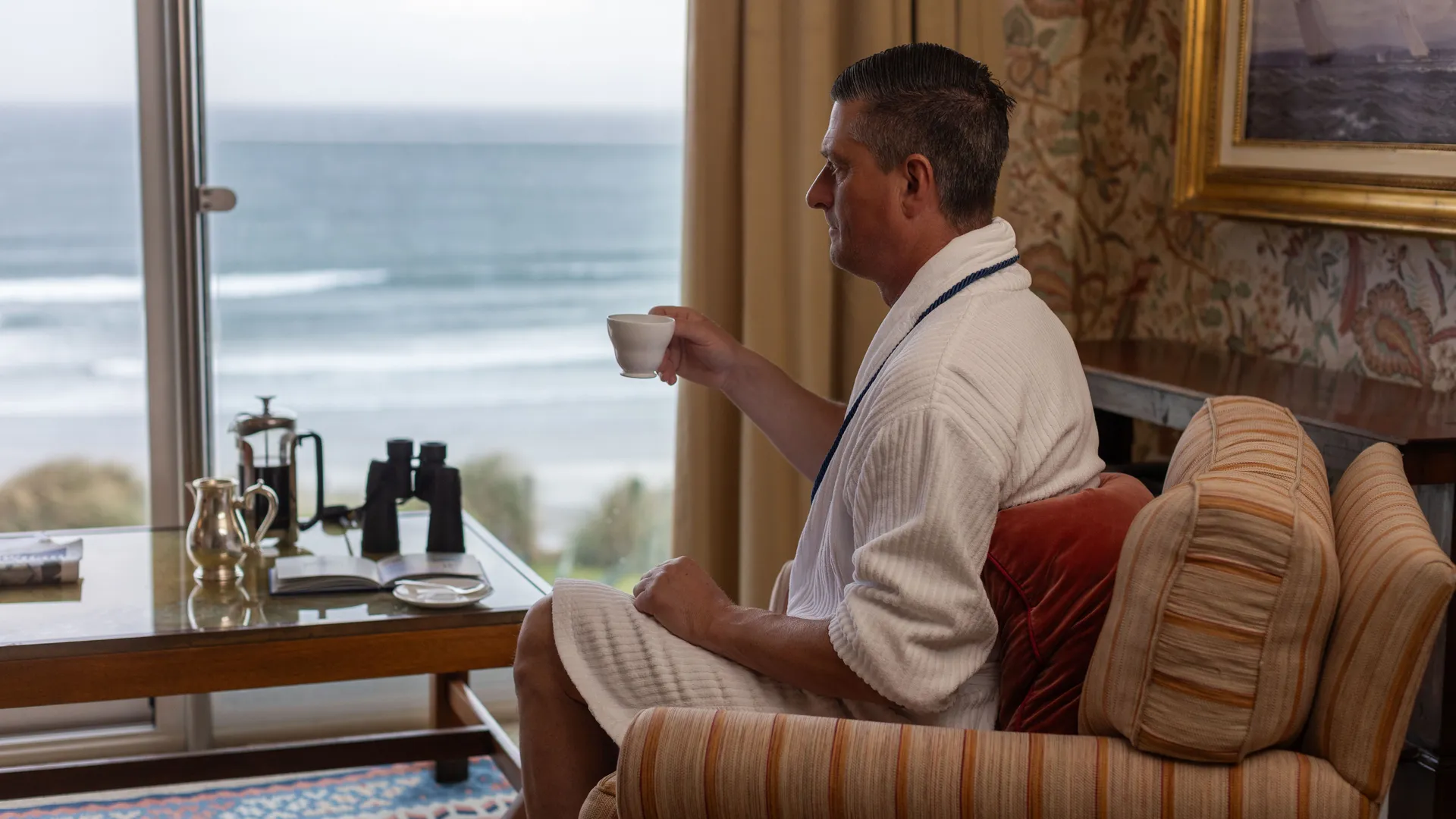 Man in a white dressing gown sitting by a seaside window, holding a cup of tea in a cosy hotel room