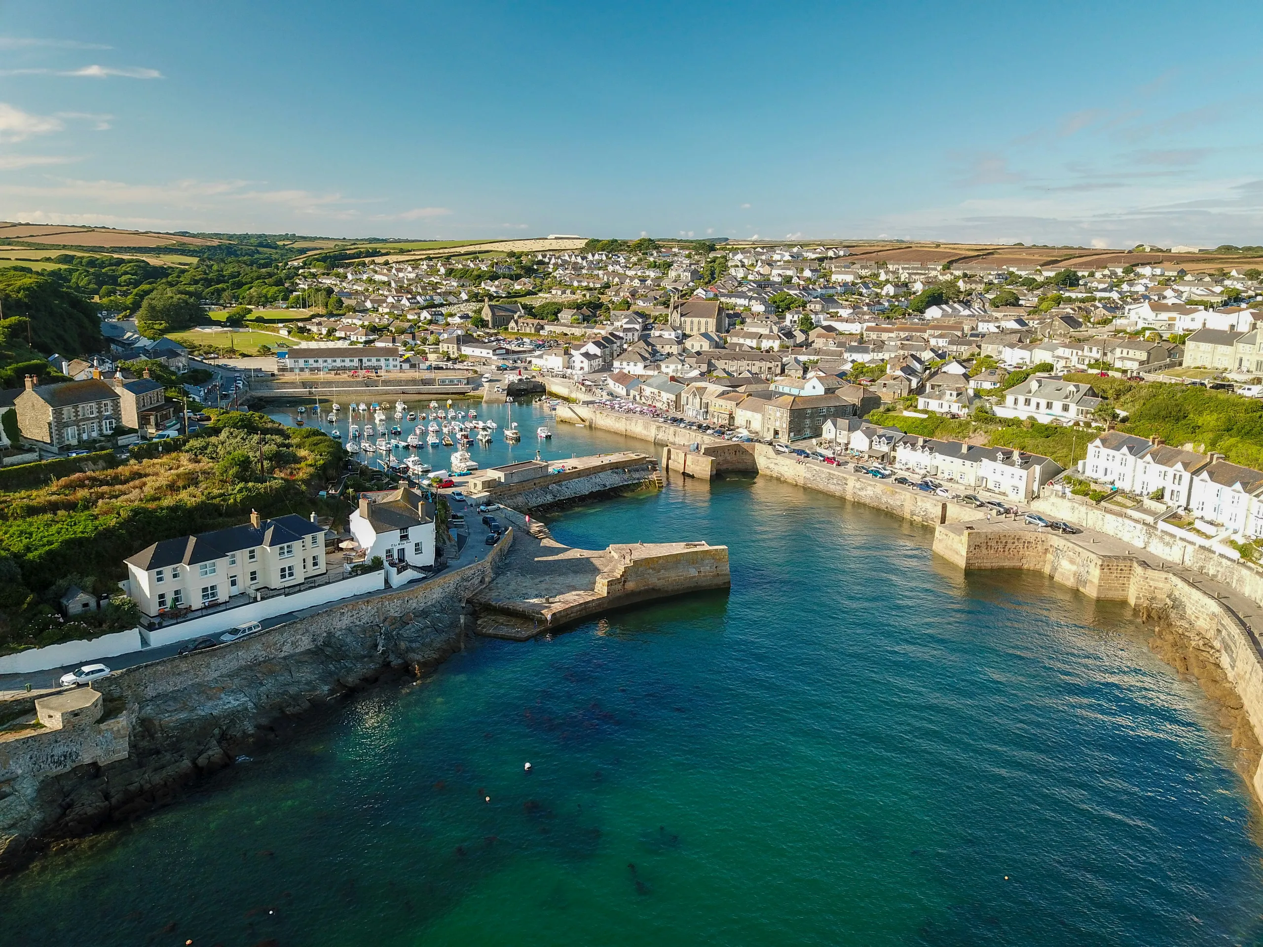 Aerial view of a coastal harbour town with stone quays, moored boats and turquoise sea under a clear blue sky.