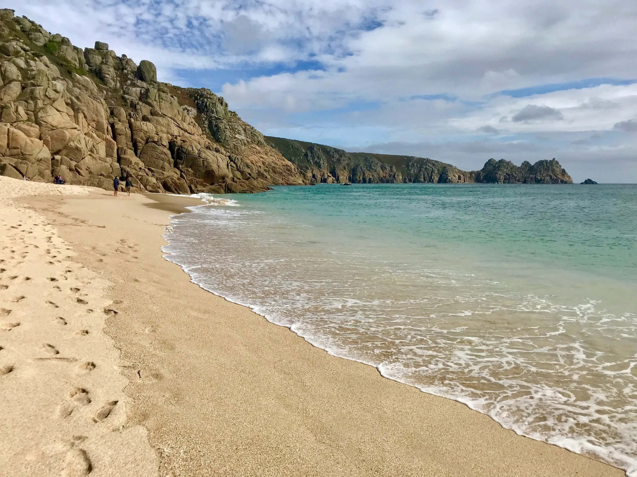 Sandy beach with gentle waves, turquoise sea and rugged rocky cliffs under a partly cloudy sky