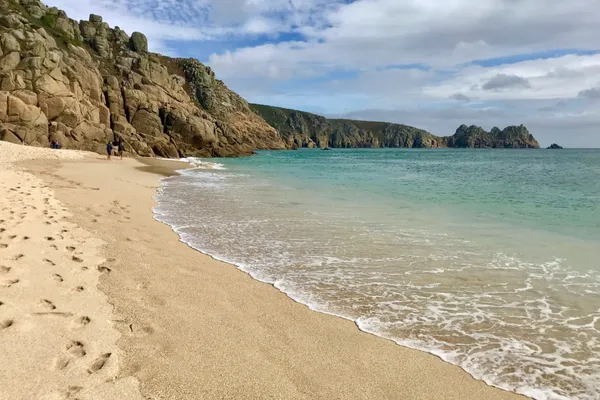 Sandy beach with gentle waves, turquoise sea and rugged rocky cliffs under a partly cloudy sky
