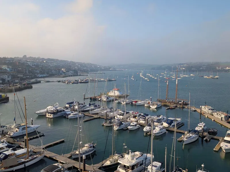 Falmouth View over a marina filled with yachts and motorboats on a calm estuary, with a hillside town and light mist in the background.
