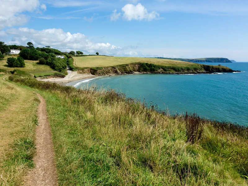 Coastal footpath at Porthcurnick Beach, Portscatho overlooking a sheltered bay with turquoise water, grassy cliffs and scattered houses under a blue sky