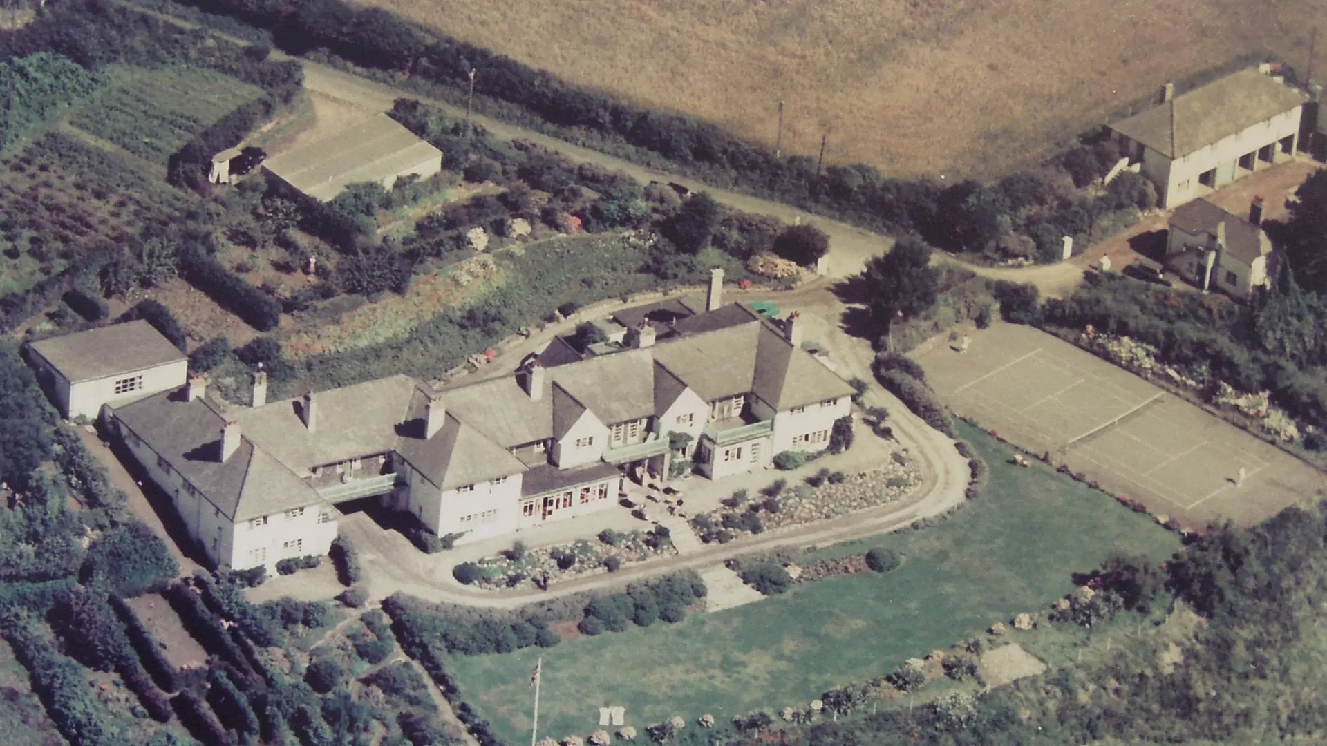 Historic aerial view of The Nare hotel and grounds on the Cornish coastline, with tennis court, gardens and surrounding countryside