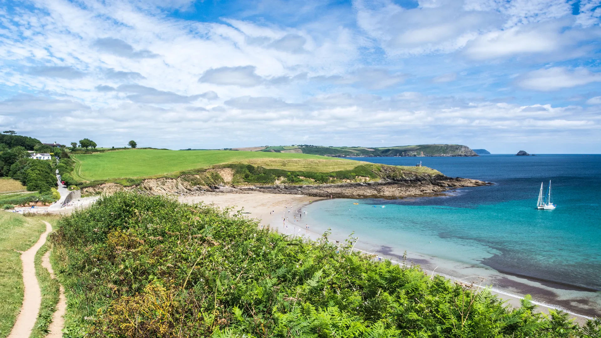 South West coastal path between Portscatho and Porthcurnick overlooking a sandy cove with turquoise water, green fields and a sailing boat under a cloudy blue sky