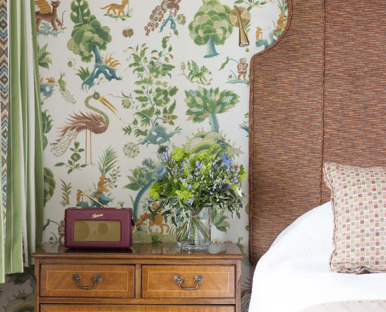 Bedside table with flowers and a vintage-style radio beside a bed with a woven headboard, set against botanical animal wallpaper and green striped curtains.