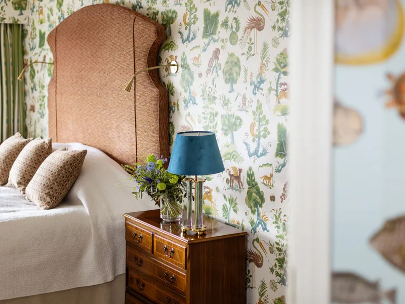 Elegant bedroom with a woven headboard, patterned wildlife wallpaper, and a wooden bedside table with a blue lamp and fresh flowers.