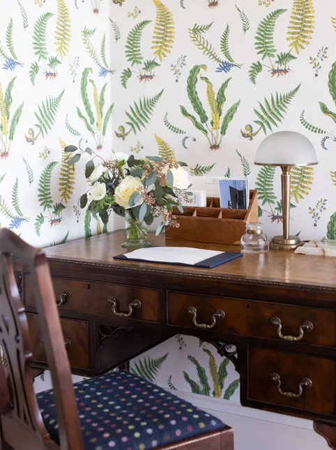 Traditional wooden writing desk with a brass table lamp and floral arrangement against botanical fern wallpaper