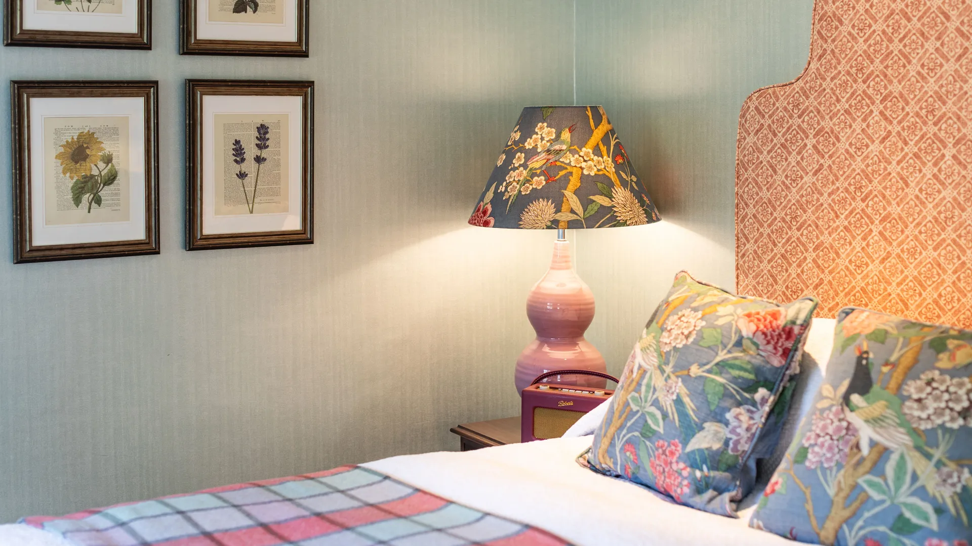Elegant bedroom with a patterned upholstered headboard, floral cushions, botanical wall art and a decorative bedside lamp.