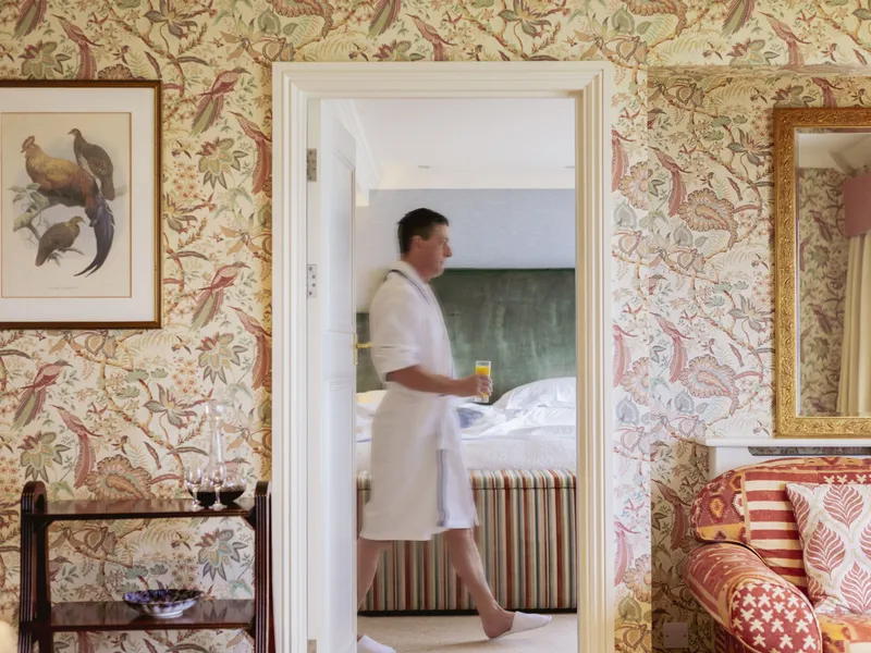 Person in a white robe walking through a doorway in an elegant bedroom with floral wallpaper and a glass of orange juice