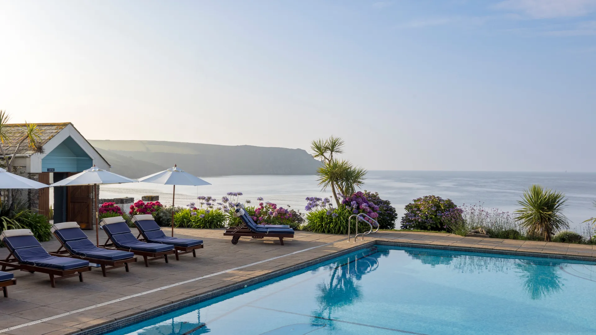 Outdoor swimming pool with sun loungers, parasols and sea views beside a coastal garden
