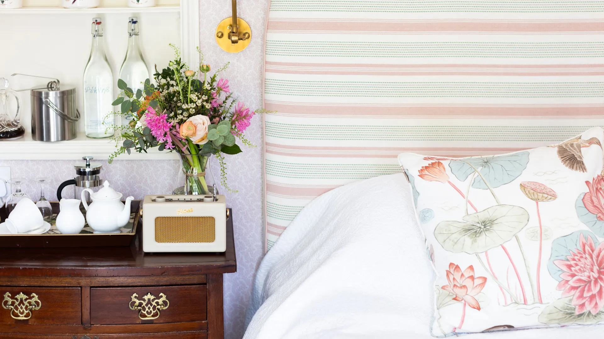 Cosy bedroom corner with a striped upholstered headboard, floral cushion, wooden bedside chest, brass wall light and fresh flowers.