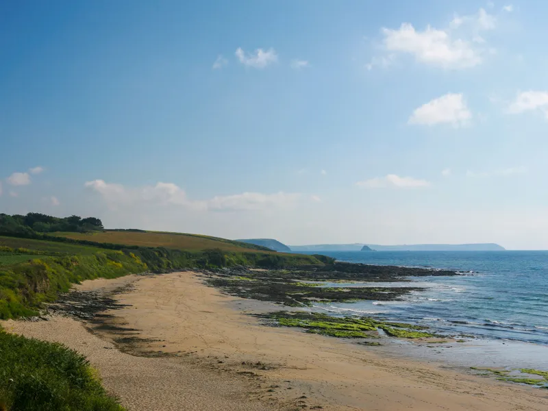 Towan Beach, St Anthony Curving sandy beach with rocky shoreline, green coastal fields and calm blue sea under a bright sky.