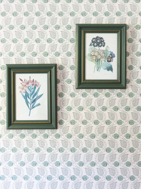 Two green-framed botanical prints hanging on a pale wallpaper with a repeating leaf and fruit pattern.