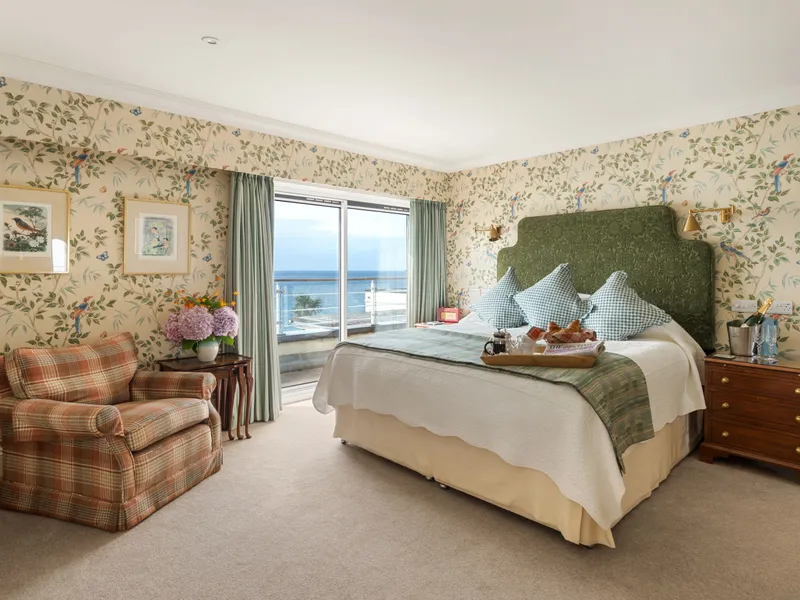Elegant bedroom with floral wallpaper, upholstered green headboard, plaid armchair and sea-view balcony doors