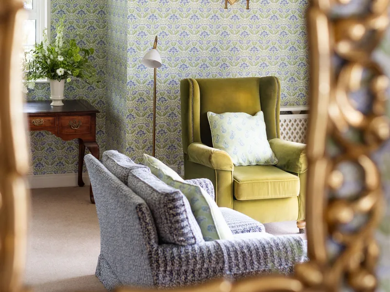 Elegant sitting room reflected in an ornate gilt mirror, with patterned wallpaper, a green armchair and blue upholstered seating.