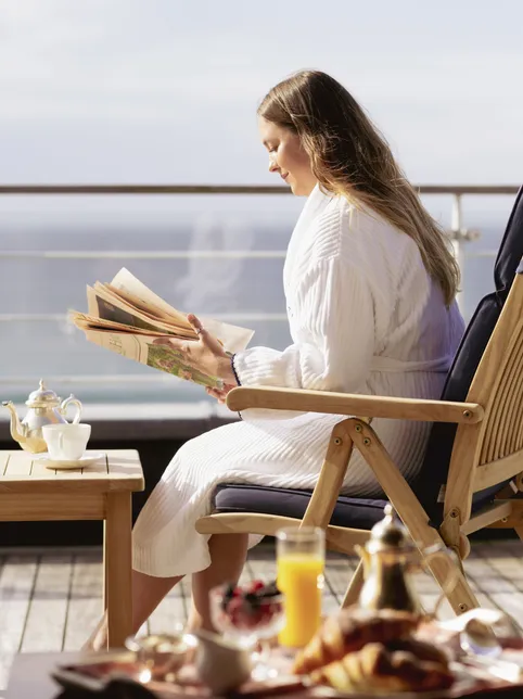 Woman in a white robe reading on a balcony with tea and breakfast overlooking the sea