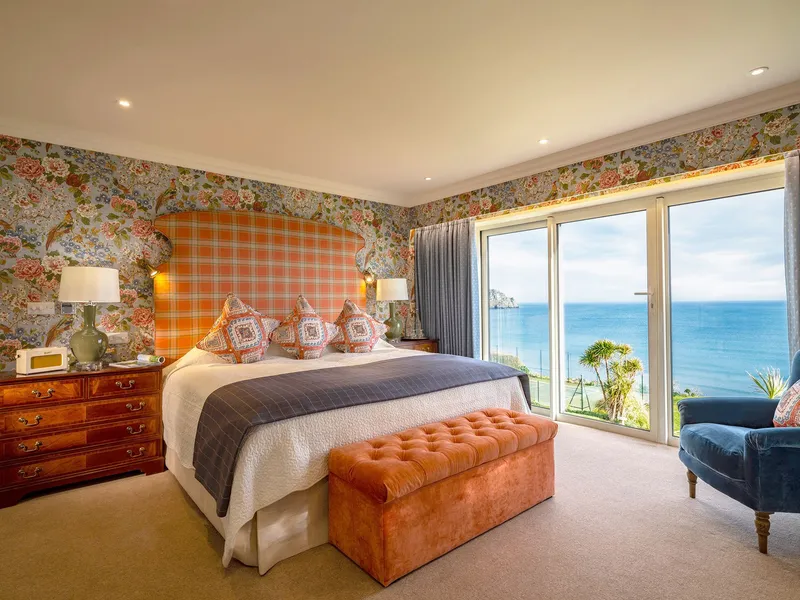 Elegant hotel suite bedroom with floral wallpaper, a large bed, and floor-to-ceiling doors opening to sea views.