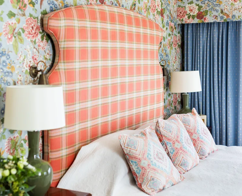 Bedroom with a coral plaid upholstered headboard, floral wallpaper, blue curtains and patterned cushions.