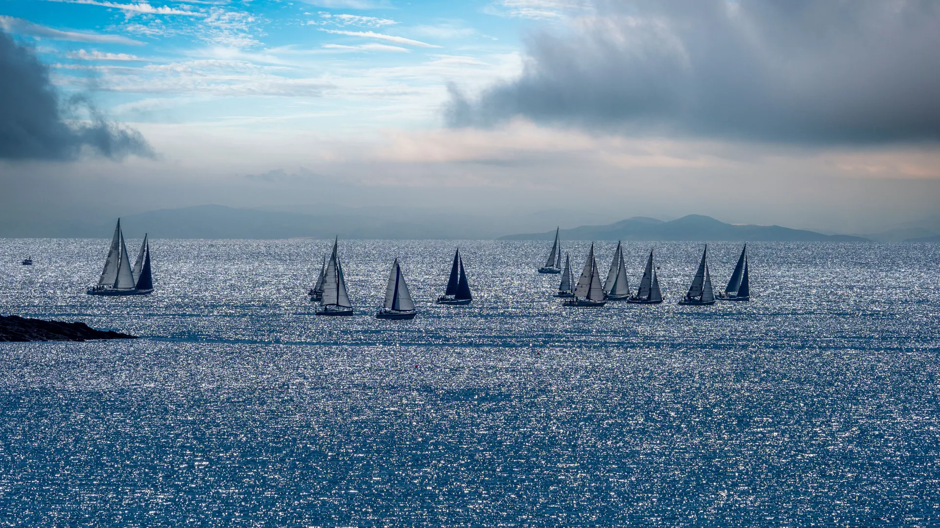 A group of sailing boats gliding across a sparkling sea beneath dramatic clouds, with distant hills on the horizon.