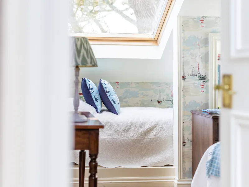 Bright attic bedroom with a skylight, white bedspread, blue cushions and nautical wallpaper.