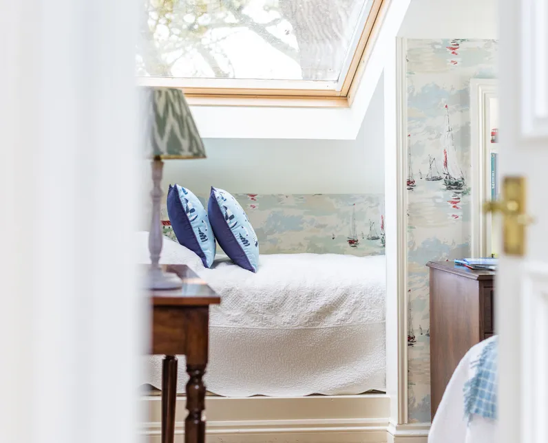 Bright attic bedroom with a skylight, white bedspread, blue cushions and nautical wallpaper.