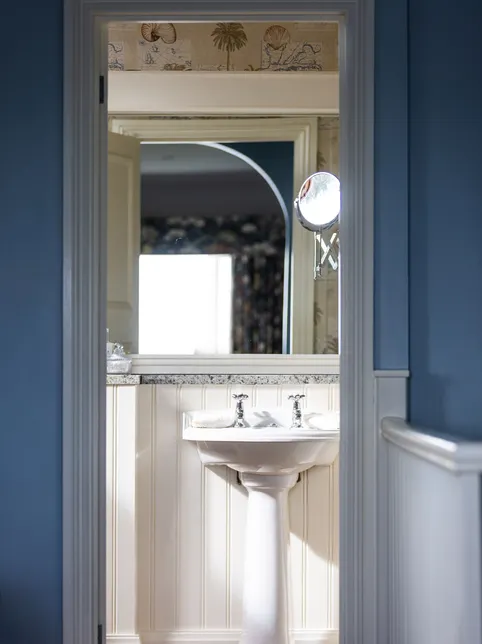 View through a doorway into a bright bathroom with a white pedestal basin, large mirror and blue walls