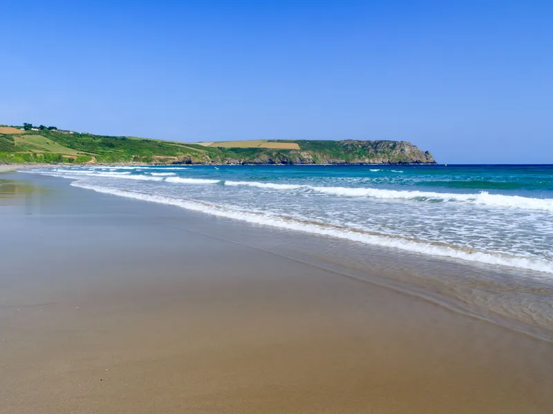Wide sandy beach with gentle waves, turquoise sea and green headlands under a clear blue sky