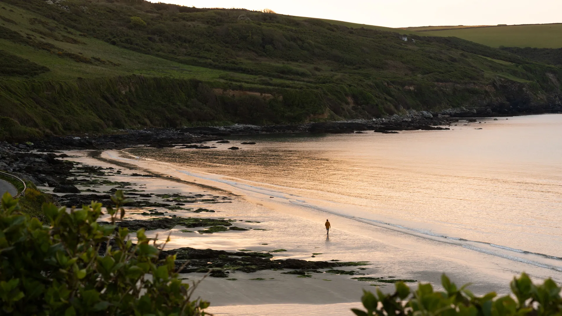 Carne Beach A solitary person walking along a quiet sandy cove at sunset, with calm sea, rocky shoreline and green cliffs in the background.