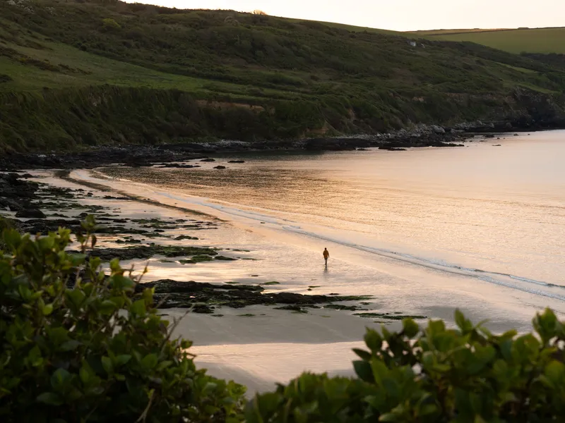 Carne Beach A solitary person walking along a quiet sandy cove at sunset, with calm sea, rocky shoreline and green cliffs in the background.