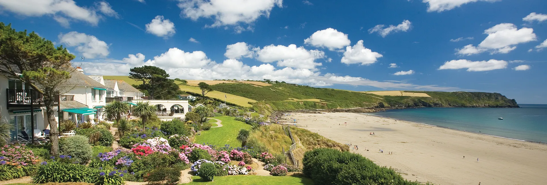 Coastal hotel and colourful gardens overlooking a sandy beach and headland under a bright blue sky